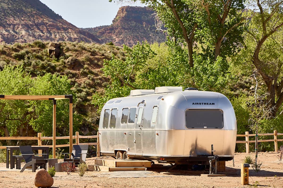 A vintage Airstream trailer with a wooden deck and outdoor seating area, set against a mountainous backdrop.