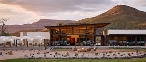 Un edificio moderno con fachada de vidrio, asientos exteriores y vistas a una montaña.