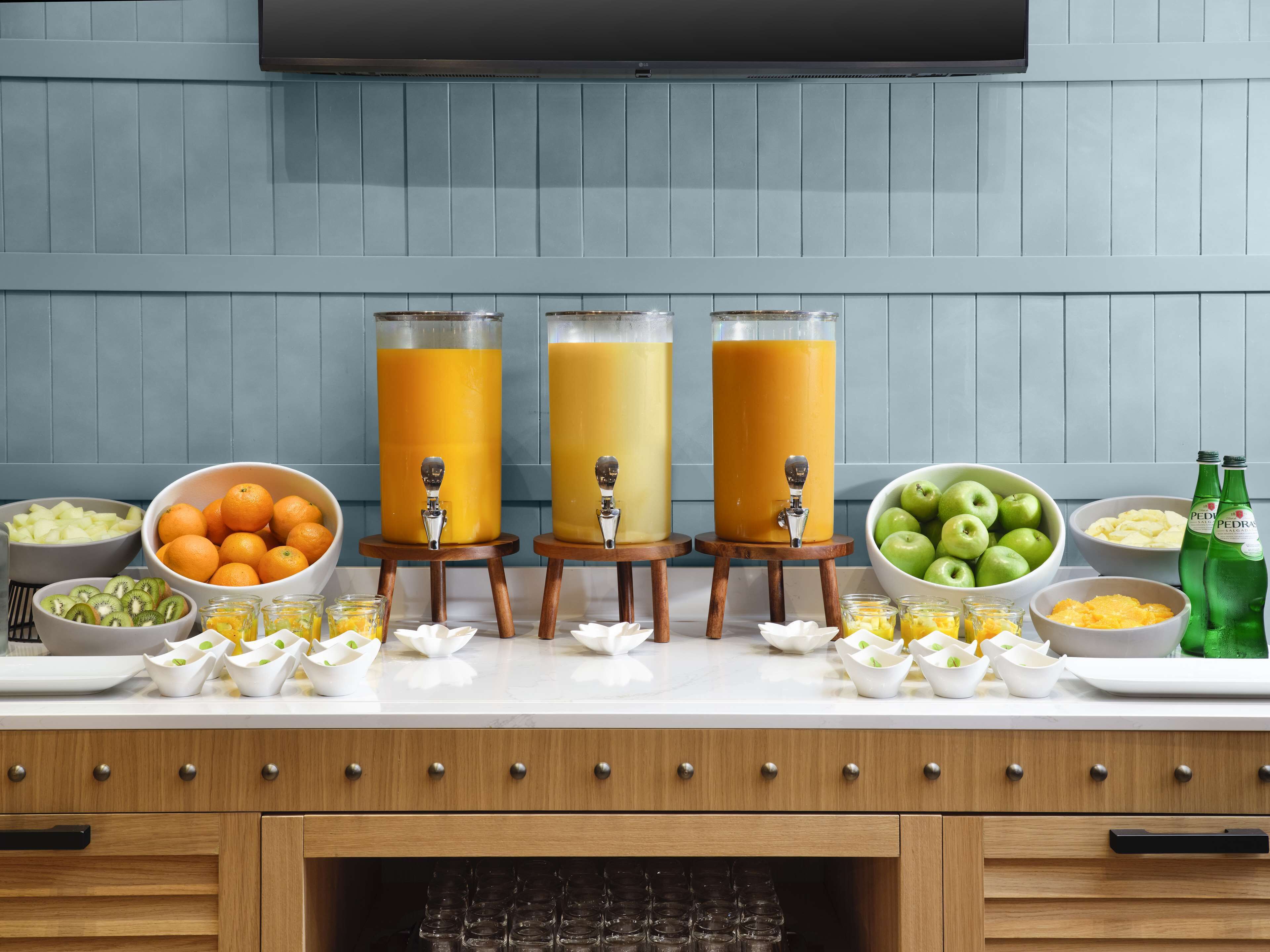 A kitchen counter with three juice dispensers, bowls of fruits, and bottles of water.