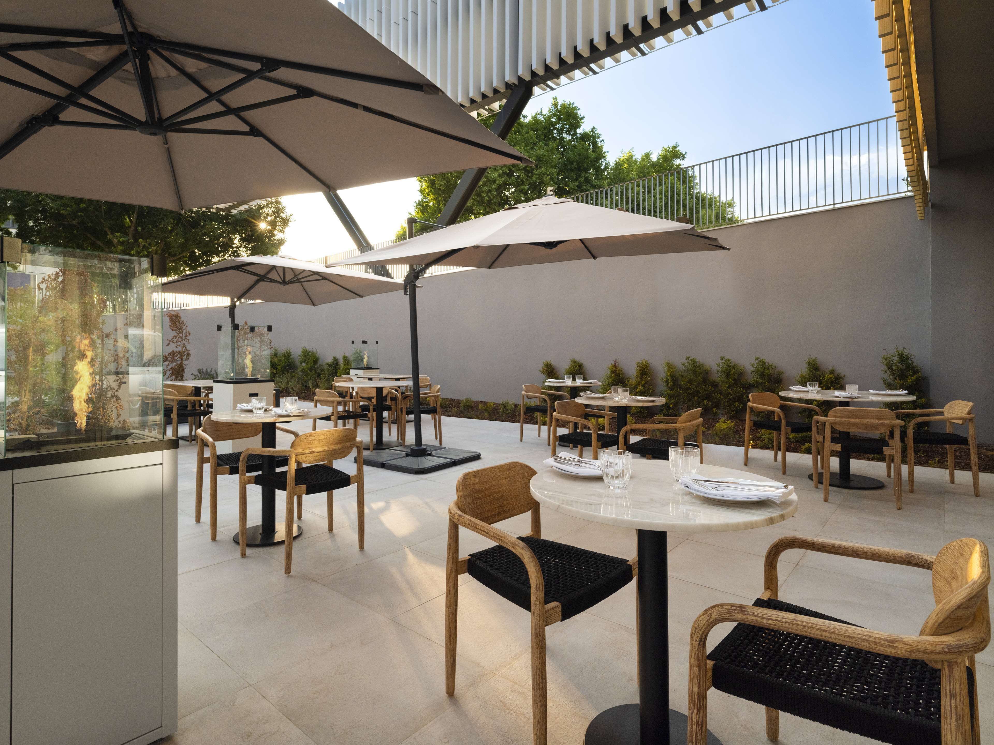 Een terras buiten met ronde tafels en stoelen, parasols en een glazen display met planten.