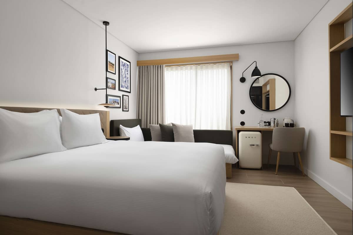 Ein modernes Hotelzimmer mit einem großen Bett, einem Schreibtisch mit Computer, einem Sessel, einer kleinen Kühlbox und einem großen Spiegel.