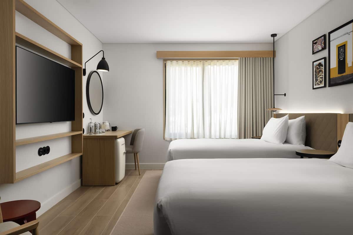 Ein modernes Hotelzimmer mit Flachbildfernseher, Schreibtisch mit Stuhl, zwei Betten und einem großen Fenster mit Vorhängen.