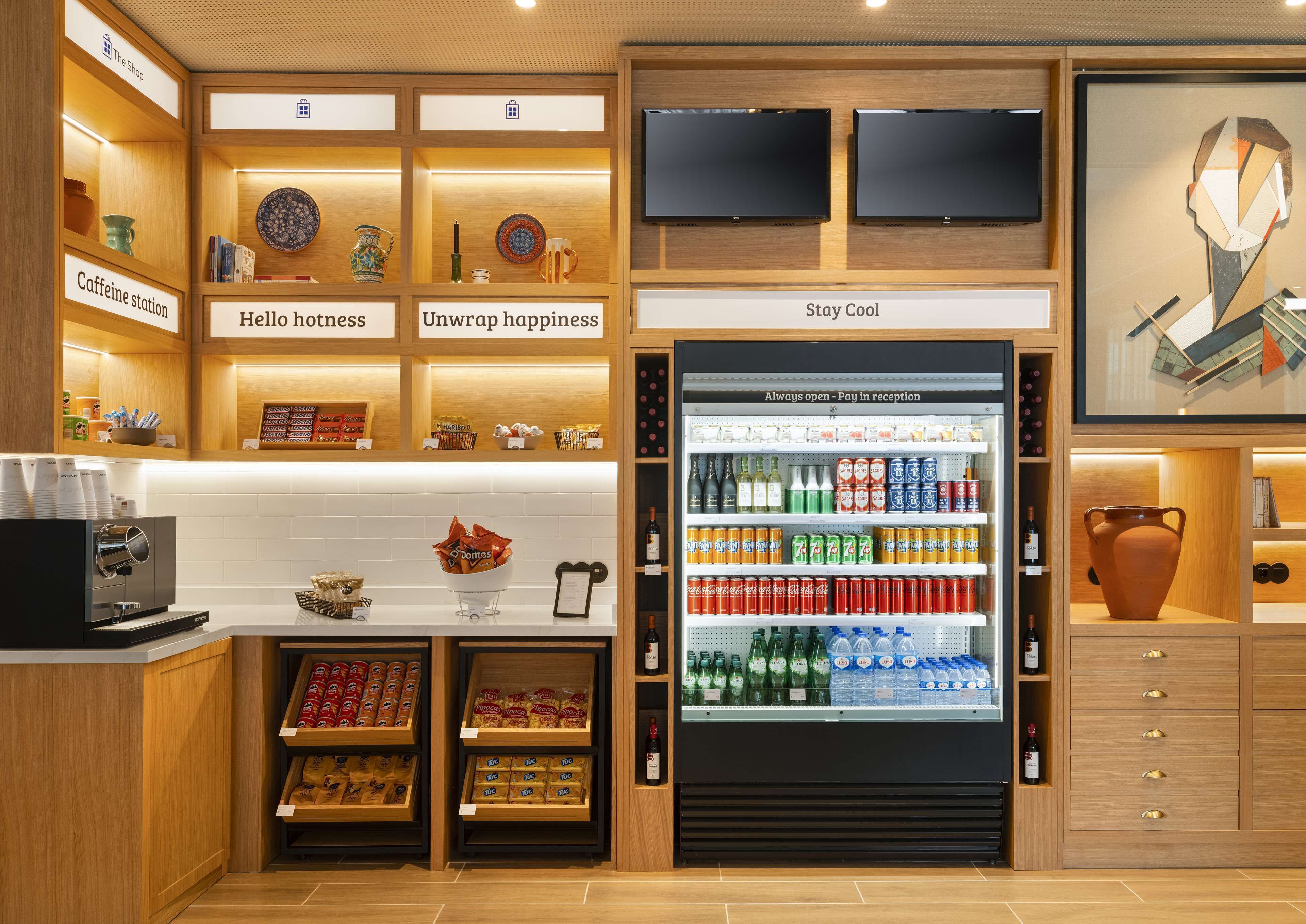 A modern hotel lobby with a coffee station, a beverage refrigerator stocked with various drinks, and a reception area.