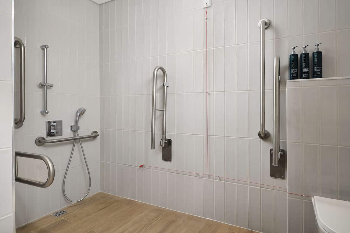 Ein modernes Badezimmer mit weißen Fliesen, Holzboden und mehreren Haltegriffen.