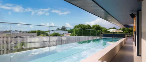 Un área de piscina moderna al aire libre con barandilla de cristal, cielo despejado y vistas de edificios y árboles circundantes.