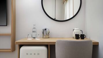 Una habitación de hotel con un escritorio de madera, un espejo redondo, un minibar, una cafetera y una silla gris.