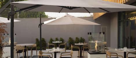 Un área de patio al aire libre con mesas y sillas redondas, cubierta por grandes sombrillas, y un expositor de vidrio con una chimenea decorativa.
