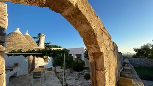 Un arco de piedra enmarca un espacio al aire libre rústico con un edificio blanco, un muro de piedra y un cielo azul despejado.