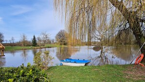 A calm lake with a small boat docked on the grassy bank, surrounded by lush greenery and trees. -  Holiday home with lake and grounds for families and groups. (L'Auvergneuse, Fenery)