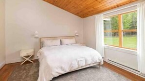 Ein Schlafzimmer mit Holzboden, einem Bett mit weißer Bettwäsche, einem Nachttisch und einem Fenster mit Blick auf Grünflächen.
