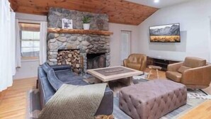 Ein Wohnzimmer mit Kamin aus Stein, Holzbalkendecke, einer Couch, einem Zweisitzer und einem Fernseher.