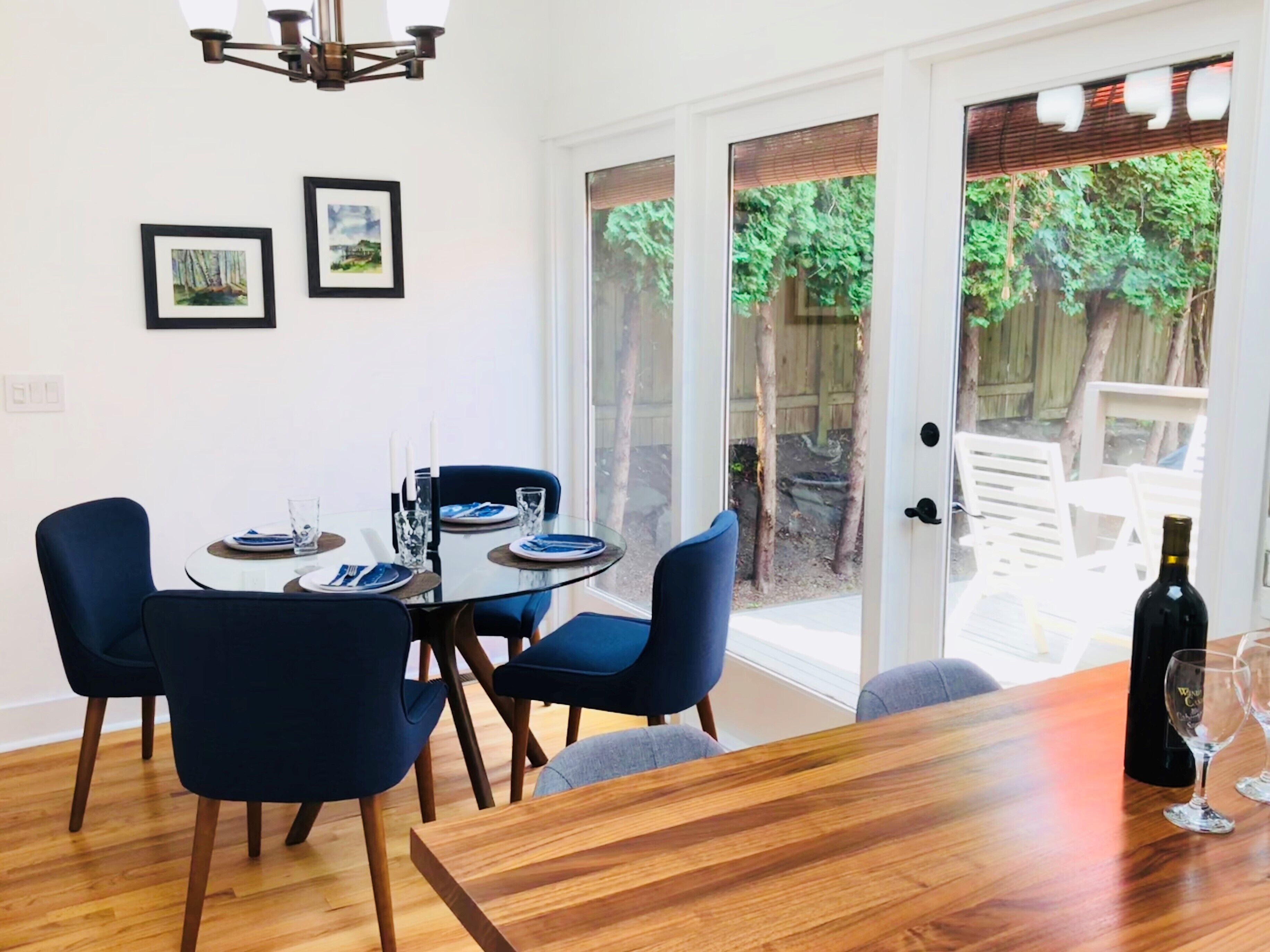 A dining area with a glass table, blue chairs, and a wooden floor. There are framed pictures on the wall and a bottle of wine on the table.