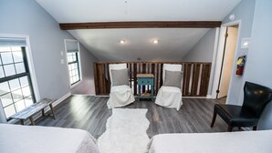 Un dormitorio estilo loft con techo inclinado, vigas de madera, un ventanal grande, dos camas blancas, un sillón de cuero negro y una pequeña mesa de madera.