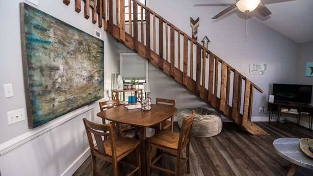 Un comedor con una mesa y sillas de madera, un cuadro abstracto grande en la pared, una escalera con barandillas de madera y un televisor en una esquina.