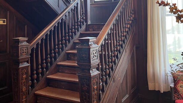Una imponente escalera con barandales de madera ornamentados y una balaustrada decorativa, que conduce a un espacio con amplios ventanales de vitral y un candelabro.