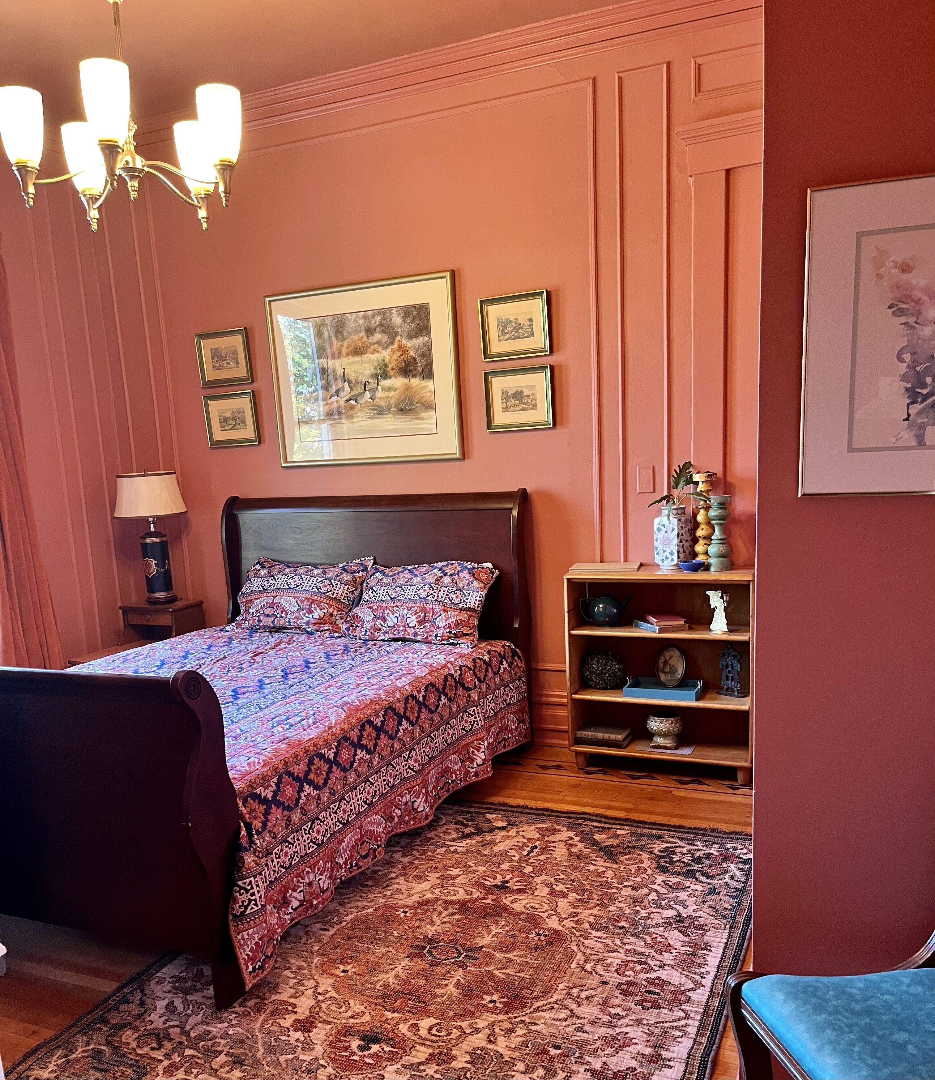 A bedroom with a patterned bedspread, a wooden nightstand with a lamp, a bookshelf with decorative items, and framed pictures on the wall.
