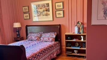 A bedroom with a patterned bedspread, a wooden nightstand with a lamp, a bookshelf with decorative items, and framed pictures on the wall.