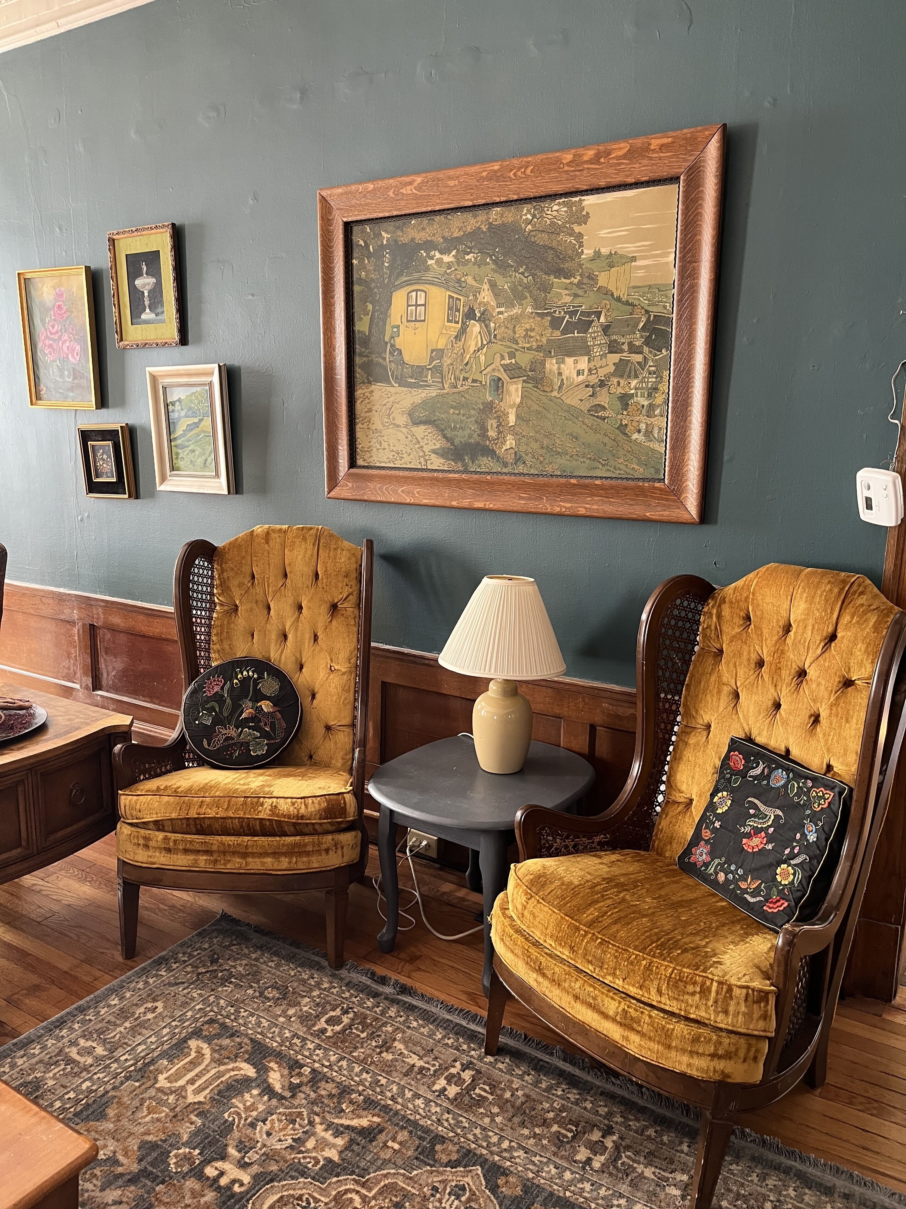 A room with two upholstered chairs, a small table with a lamp, and a large framed painting on the wall.