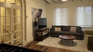 Un salón con sofá, mesa de centro y televisor. Cuenta con ventilador de techo, chimenea y ventana con persianas.