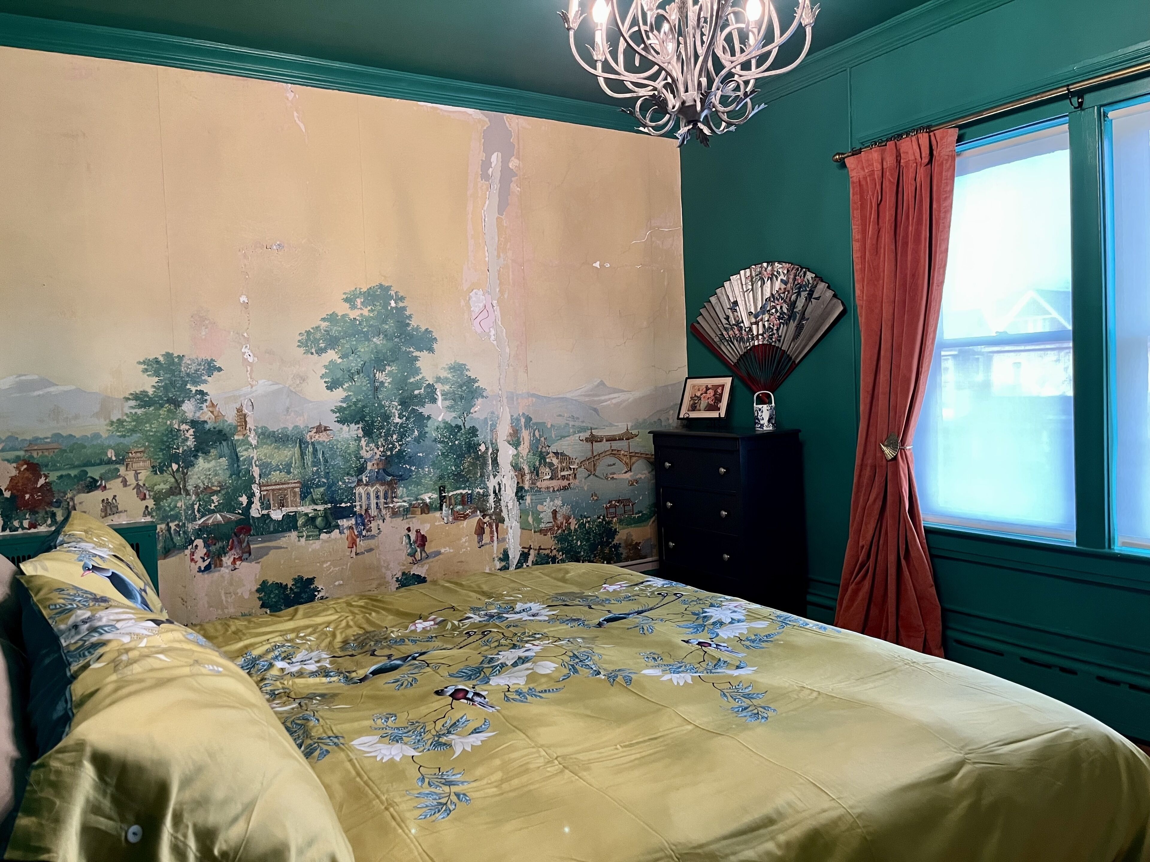 Een slaapkamer met een groot muurschildering, een bed met bloemenbedekte lakens, een dressoir en een raam met gordijnen.