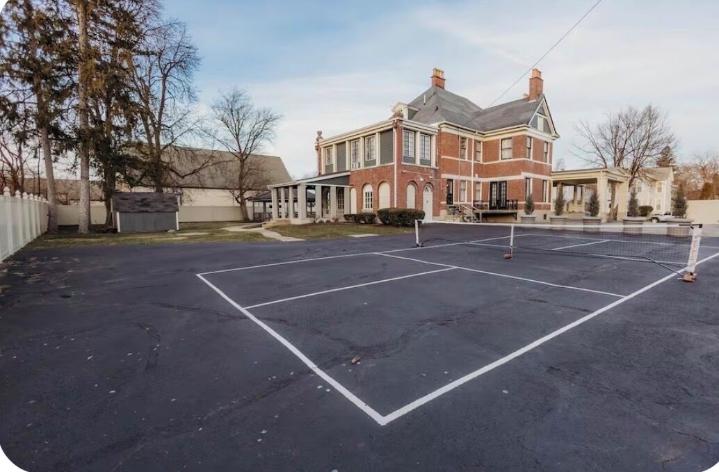 Un court de tennis avec un filet devant un grand bâtiment en briques, doté de nombreuses fenêtres et d’un porche abrité.