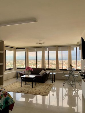 Un salon moderne comprenant un canapé, une table basse et un coin repas, de grandes fenêtres offrant une vue sur la ville et un ventilateur de plafond.