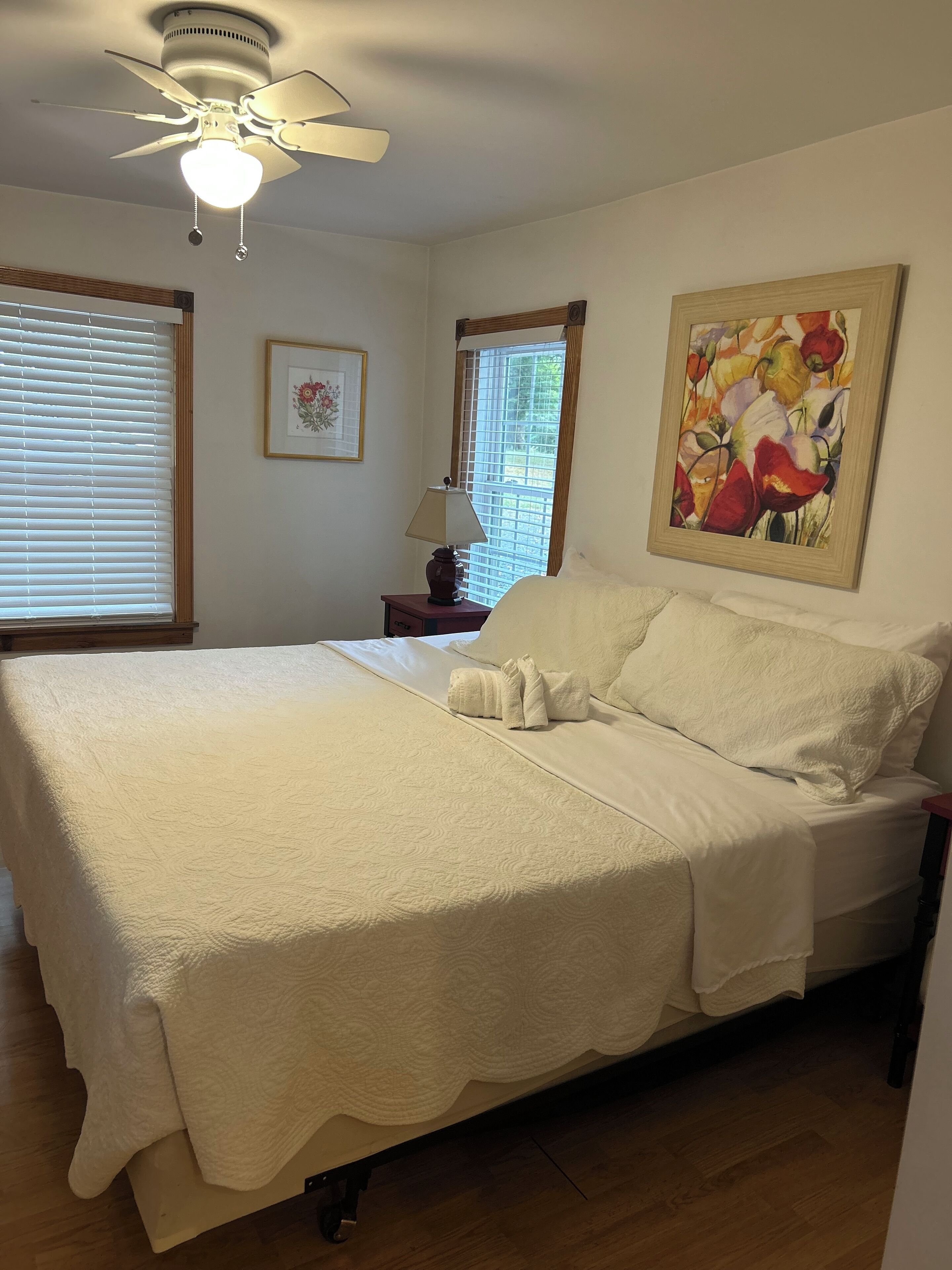 A neatly made bed with a white comforter and pillows, a ceiling fan, a window with blinds, a bedside table with a lamp, and a framed floral painting on the wall.
