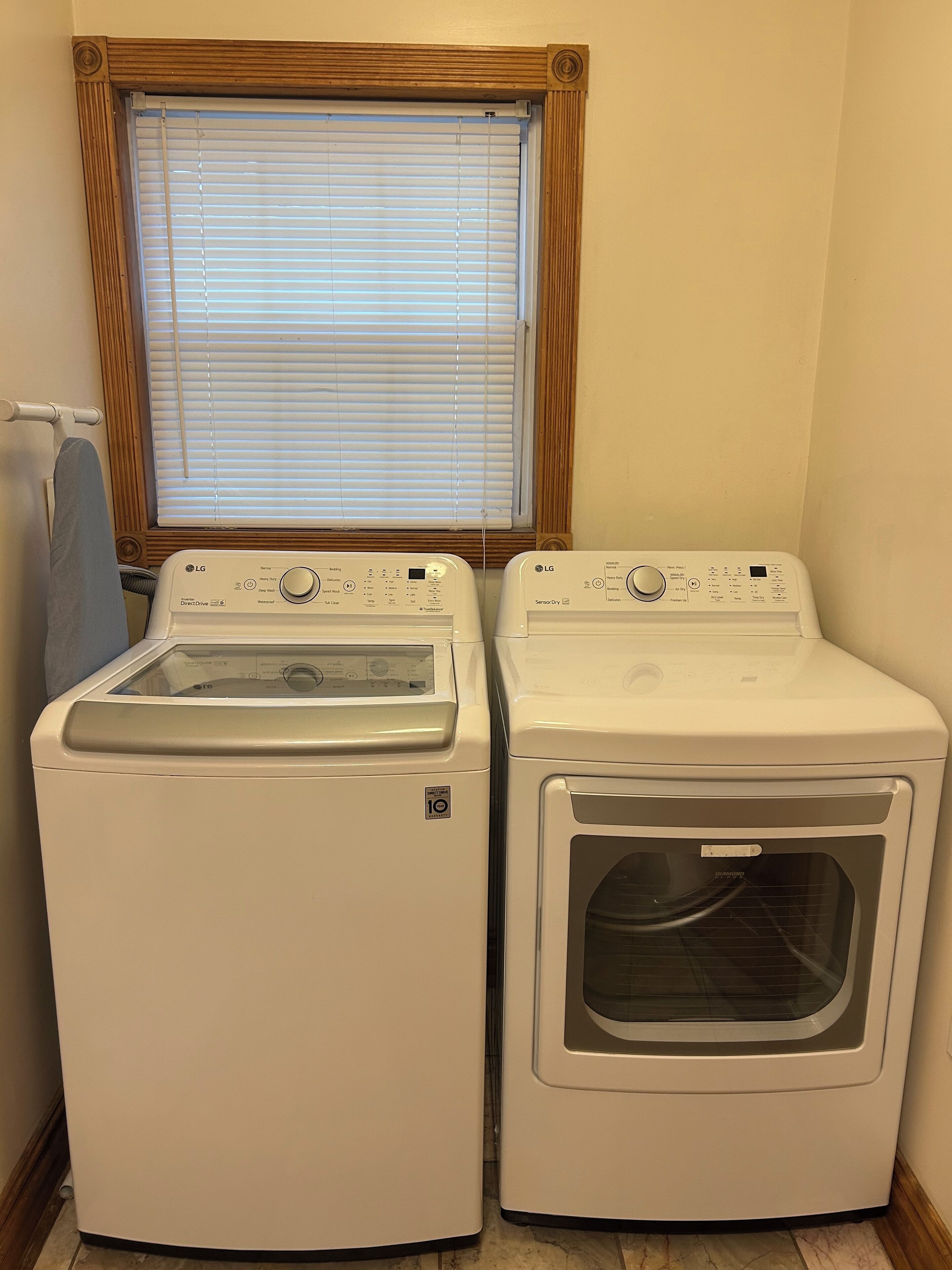 A laundry room with two white front-loading washers and dryers, a window with blinds, and a towel rack.
