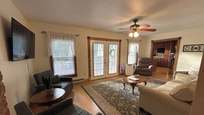 A living room with a sofa, armchairs, a coffee table, and a TV. There are two windows with blinds, a ceiling fan, and a door leading to a kitchen. - Step into history & unwind at Elixir apt a charming 🏡 at the❤️of NYHudson Valley (Highland)
