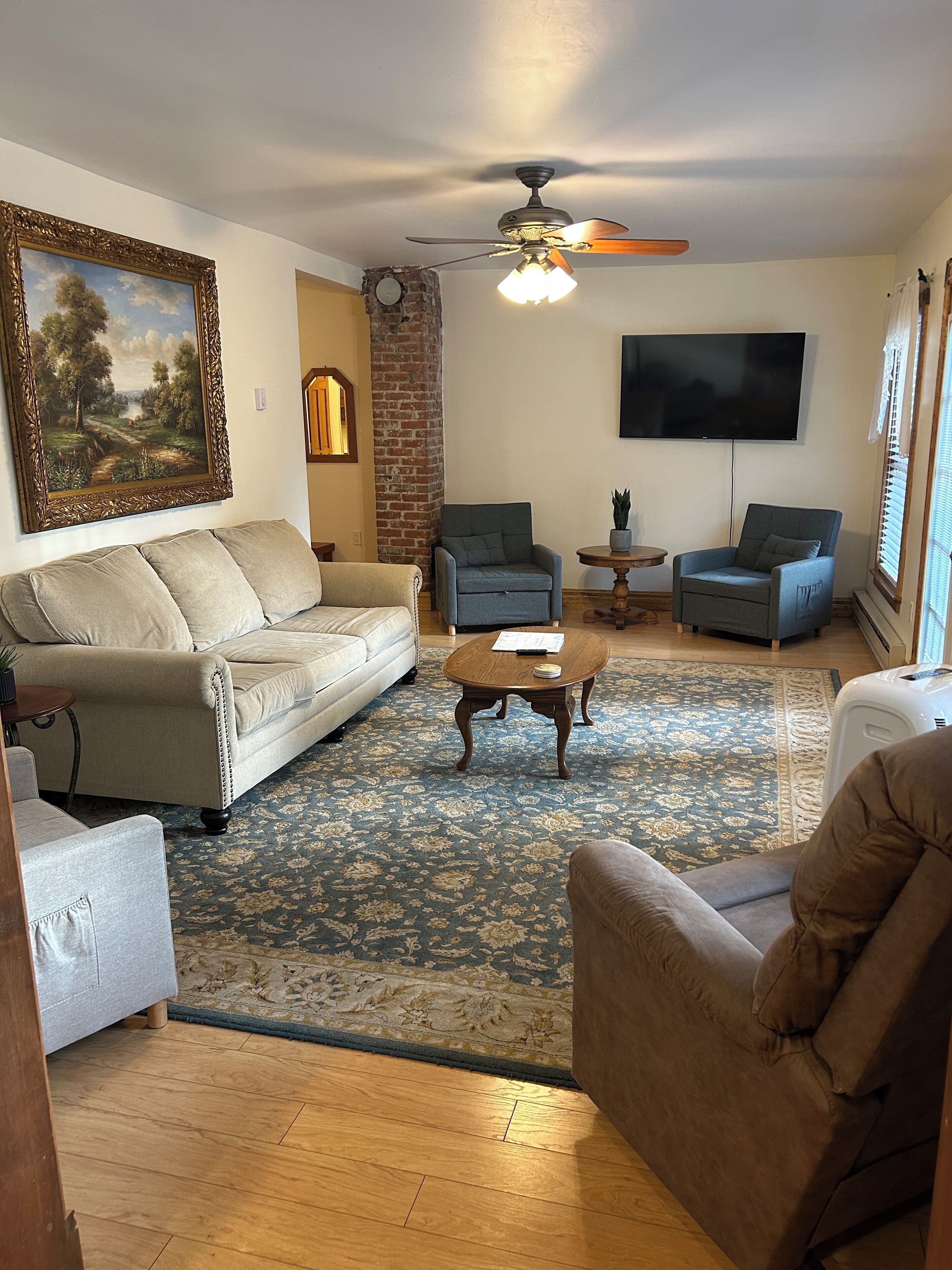 Un salon avec un canapé beige, deux fauteuils, une table basse, un tapis à motifs, un ventilateur de plafond, un téléviseur et un tableau accroché au mur.
