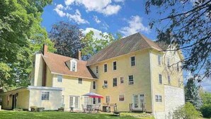 A large, multi-story yellow building with a prominent chimney, a covered patio area, and a well-maintained lawn. - Step into history & unwind at Elixir apt a charming 🏡 at the❤️of NYHudson Valley (Highland)