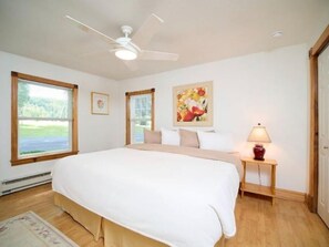 Quarto com uma cama grande, um ventilador de teto, duas janelas, uma mesa de cabeceira com abajur e um quadro na parede.