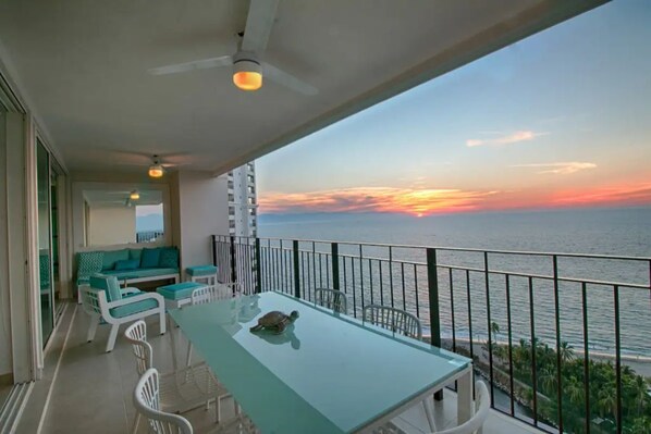 A balcony with a glass table and chairs, a turtle on the table, and a sunset view over the ocean.