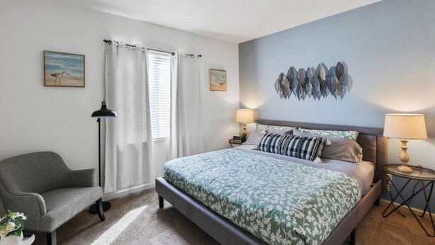 Un dormitorio con cama, dos mesitas de noche, una silla, una lámpara de pie y una pared con estampado de hojas.