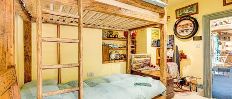 Een eenvoudige kamer met een houten stapelbed, een klein bureau met stoel, en diverse verzamelobjecten op planken.