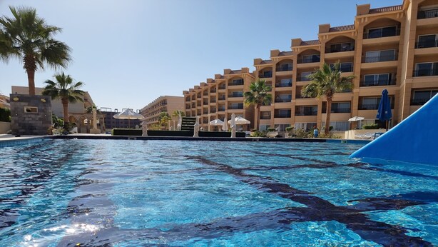 Une piscine à l’eau cristalline, entourée de palmiers et bordée d’un hôtel à plusieurs étages doté de balcons.