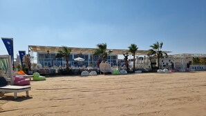 Un espace aménagé au bord de la plage, avec des chaises longues et des fauteuils en forme de poire, un coin salon abrité et un bâtiment doté de grandes fenêtres et bordé de palmiers.