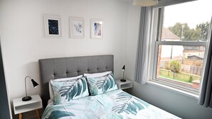 Ein Schlafzimmer mit grauem, bezogenem Kopfteil, einem Bett mit einem Bettbezug mit Blättermotiv, zwei Nachttischen mit Lampen und gerahmten Kunstwerken an der Wand.