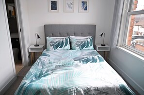 A bedroom with a grey upholstered headboard, a bed with a leaf-patterned bedspread, two bedside tables with lamps, and three framed artworks on the wall.