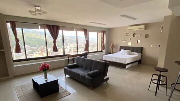 Ruime hotelkamer met een groot bed, een sofa, een kleine tafel met een bloevas en uitzicht op de stad door grote ramen.