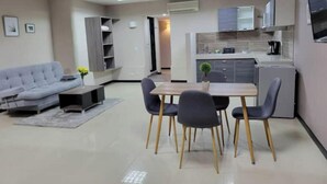 Uma sala de estar moderna com sofá, mesa de jantar com cadeiras, pequena cozinha com armários e eletrodomésticos, e um corredor que dá para outro cômodo.