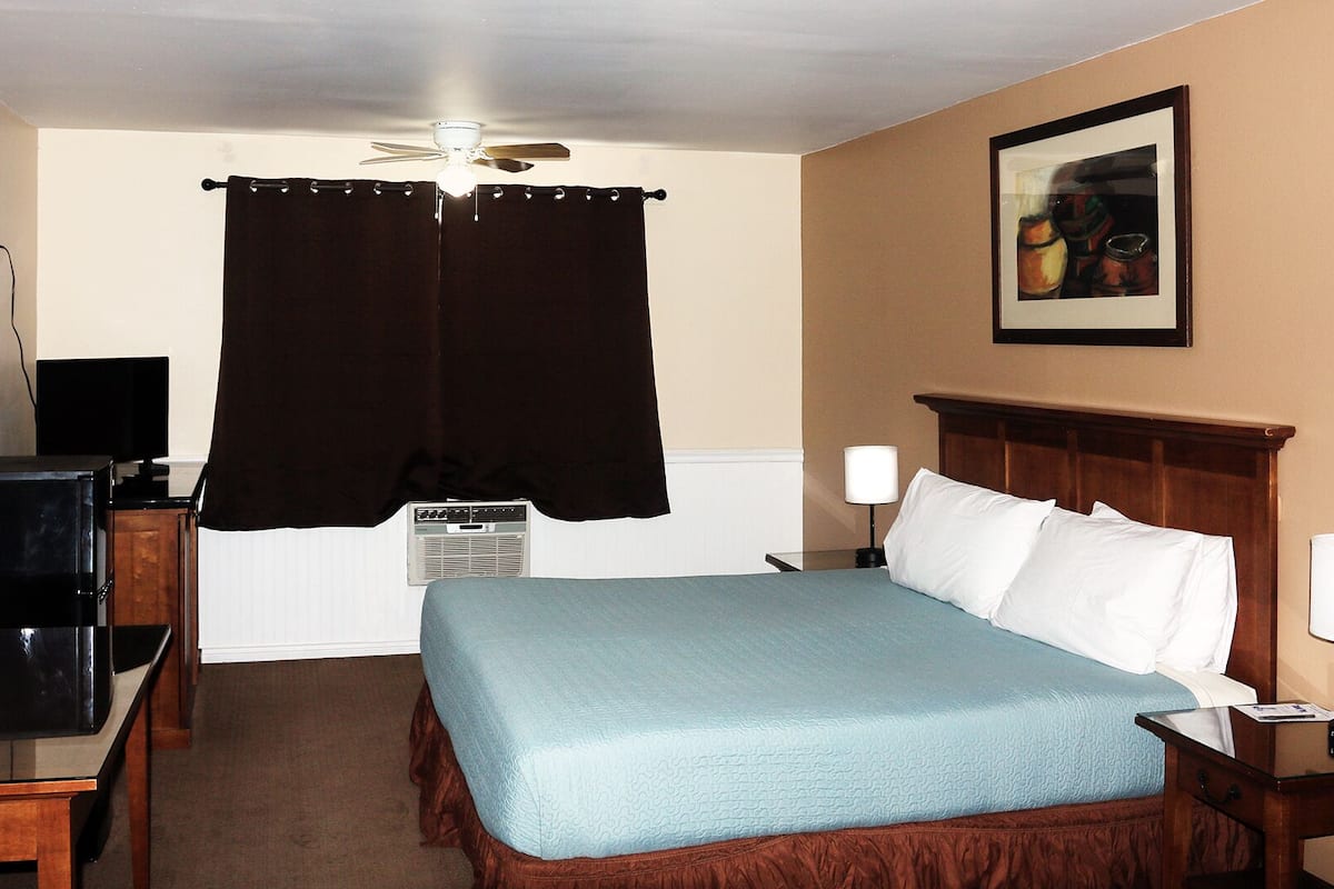 A hotel room with a bed, bedside tables, a desk with a computer, a ceiling fan, and a painting on the wall.