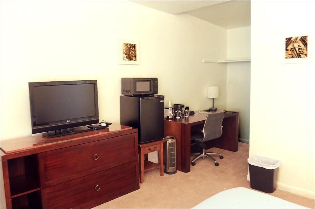 A hotel room with a wooden dresser, a television, a microwave, a desk with a chair, a lamp, and a trash bin.