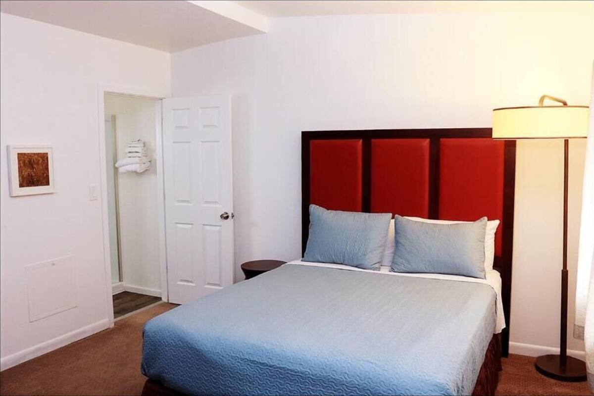 A hotel room with a bed, a nightstand, a lamp, a door, and a wall-mounted artwork.