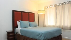 Ein ordentlich bezogenes Bett mit braunem Kopfteil, einer Nachttischlampe und einem Fenster mit durchscheinenden Vorhängen.