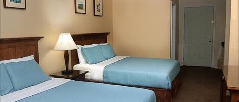 A hotel room with two beds, a nightstand with a lamp, and a door.