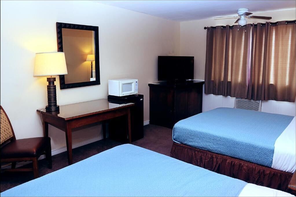 A hotel room with two beds, a desk with a lamp, a microwave, a television, and a ceiling fan.