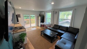 A spacious living room with a black leather sofa, a coffee table, and a TV mounted on the wall. There is a sliding glass door leading to a balcony with a wooden deck.