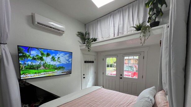 A bedroom with a bed, a TV on the wall, an air conditioner, and a door with windows.