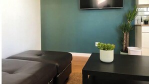 A modern living room with a teal accent wall, a black leather sofa, a small black table with a white vase, and a flat-screen TV mounted above the sofa.
